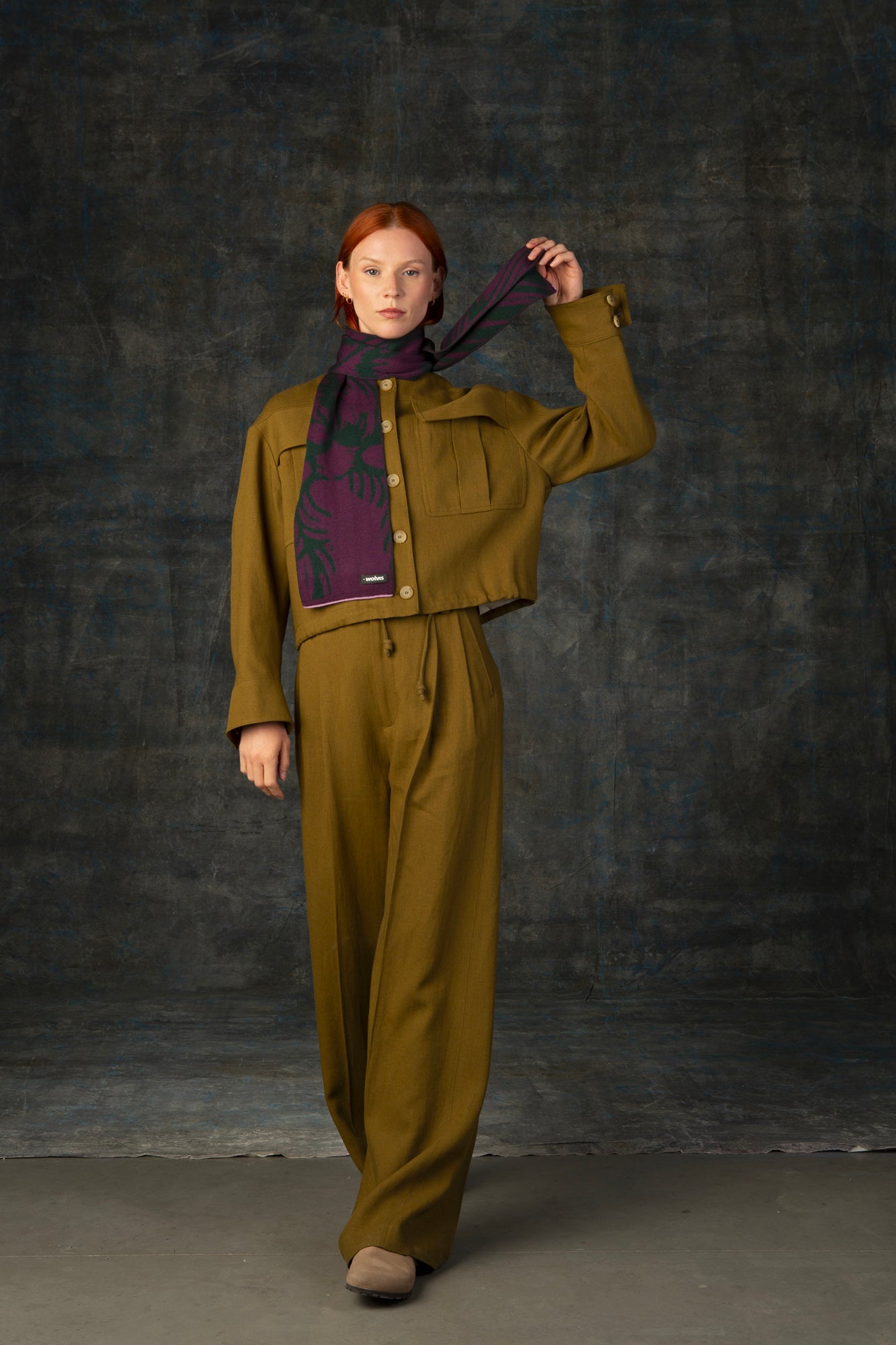 Female model wearing an olive green outfit throwing a purple and forest green mini scarf, with nature-inspired pattern, around her neck.