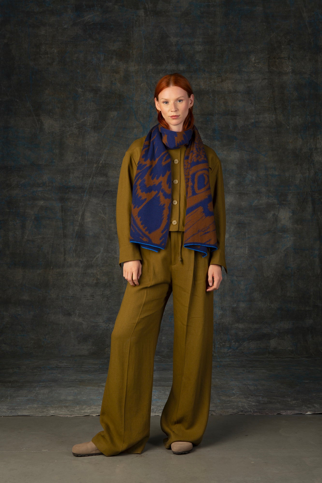 Female model wearing an olive green outfit with a brown and blue maxi scarf, with an organic pattern, draped around her neck.