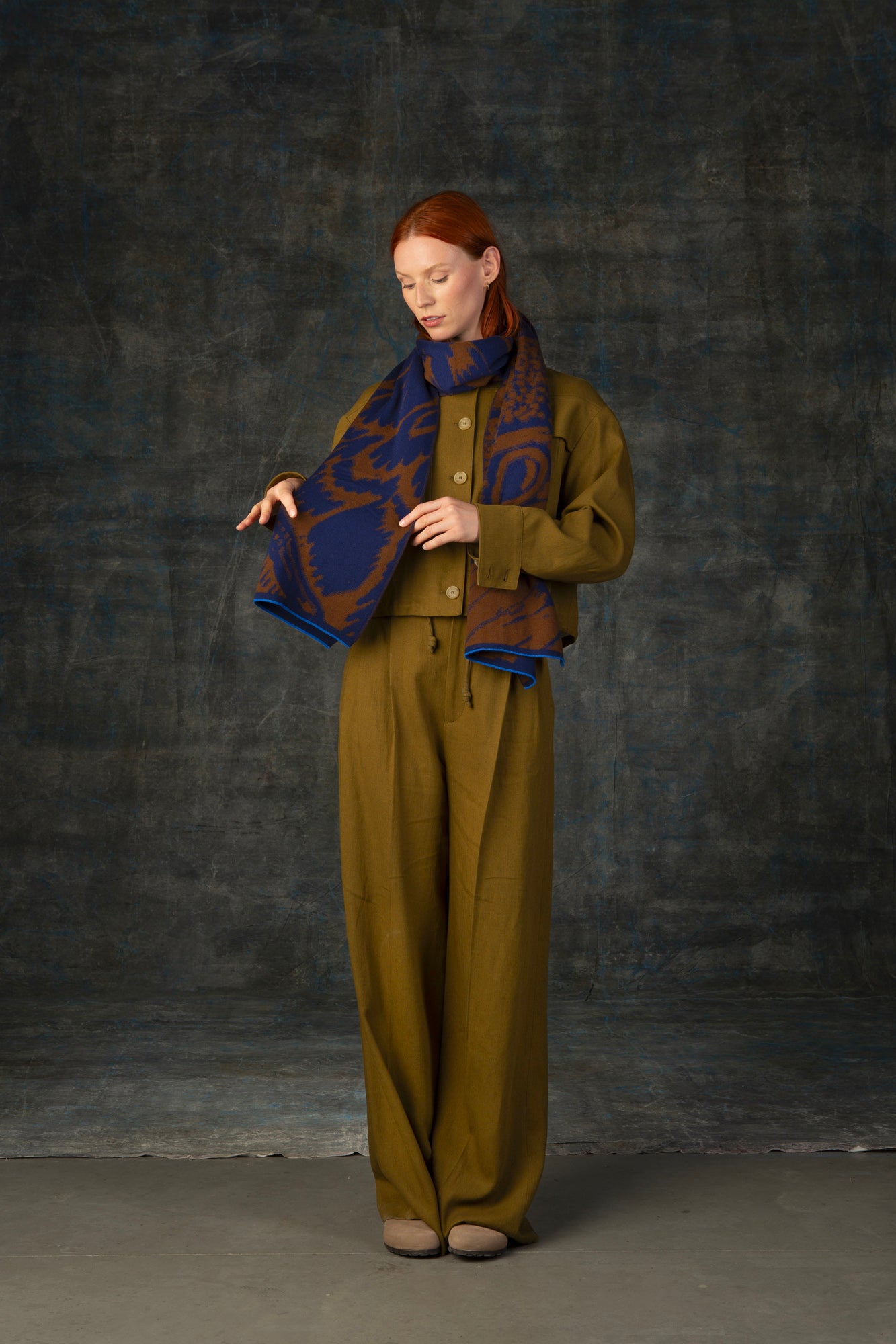 Female model wearing an olive green outfit with a brown and blue maxi scarf, with an organic pattern, draped around his neck.