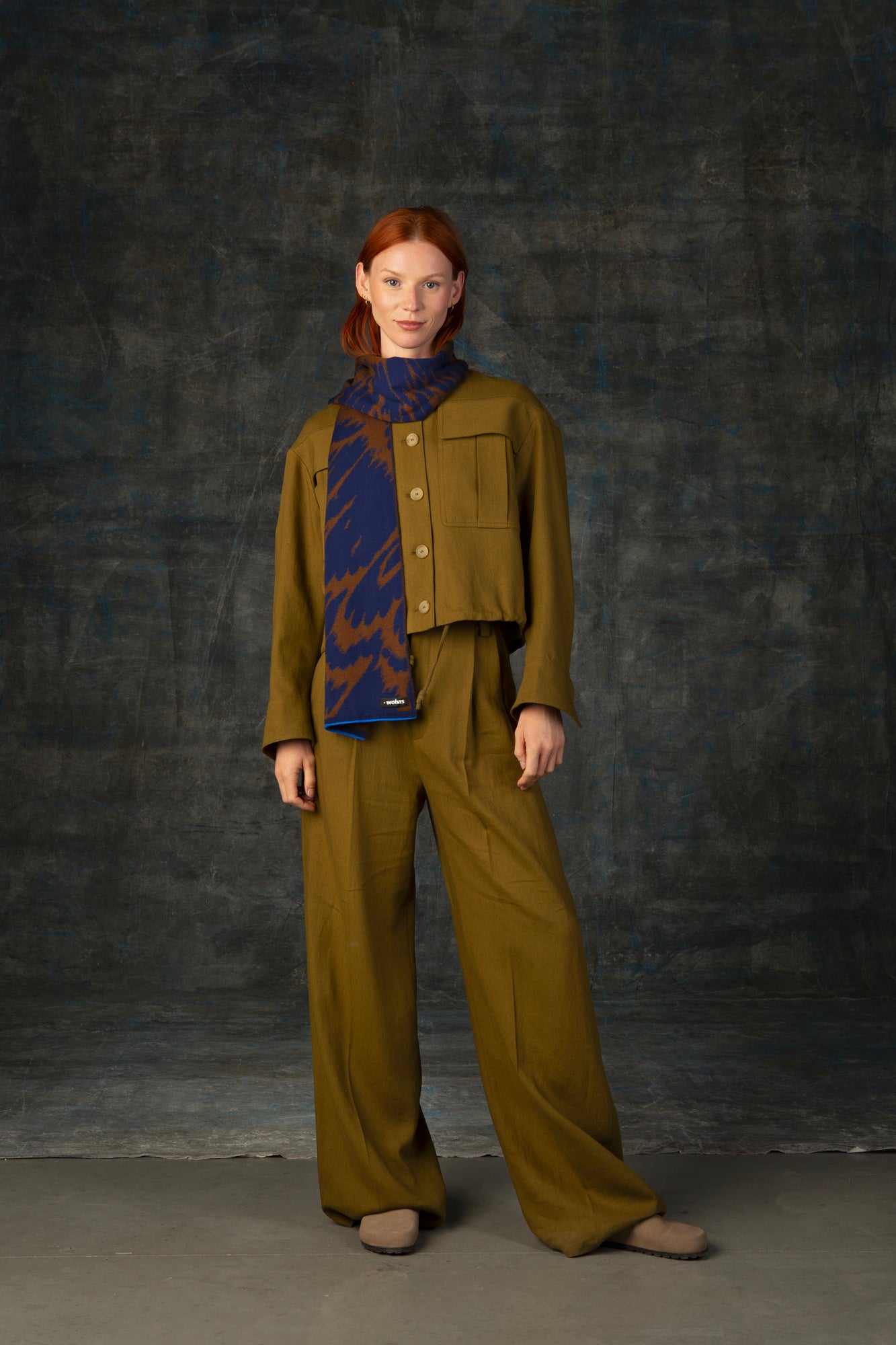 Female model wearing an olive green outfit with a brown and blue midi scarf, with an organic pattern, draped around her neck.