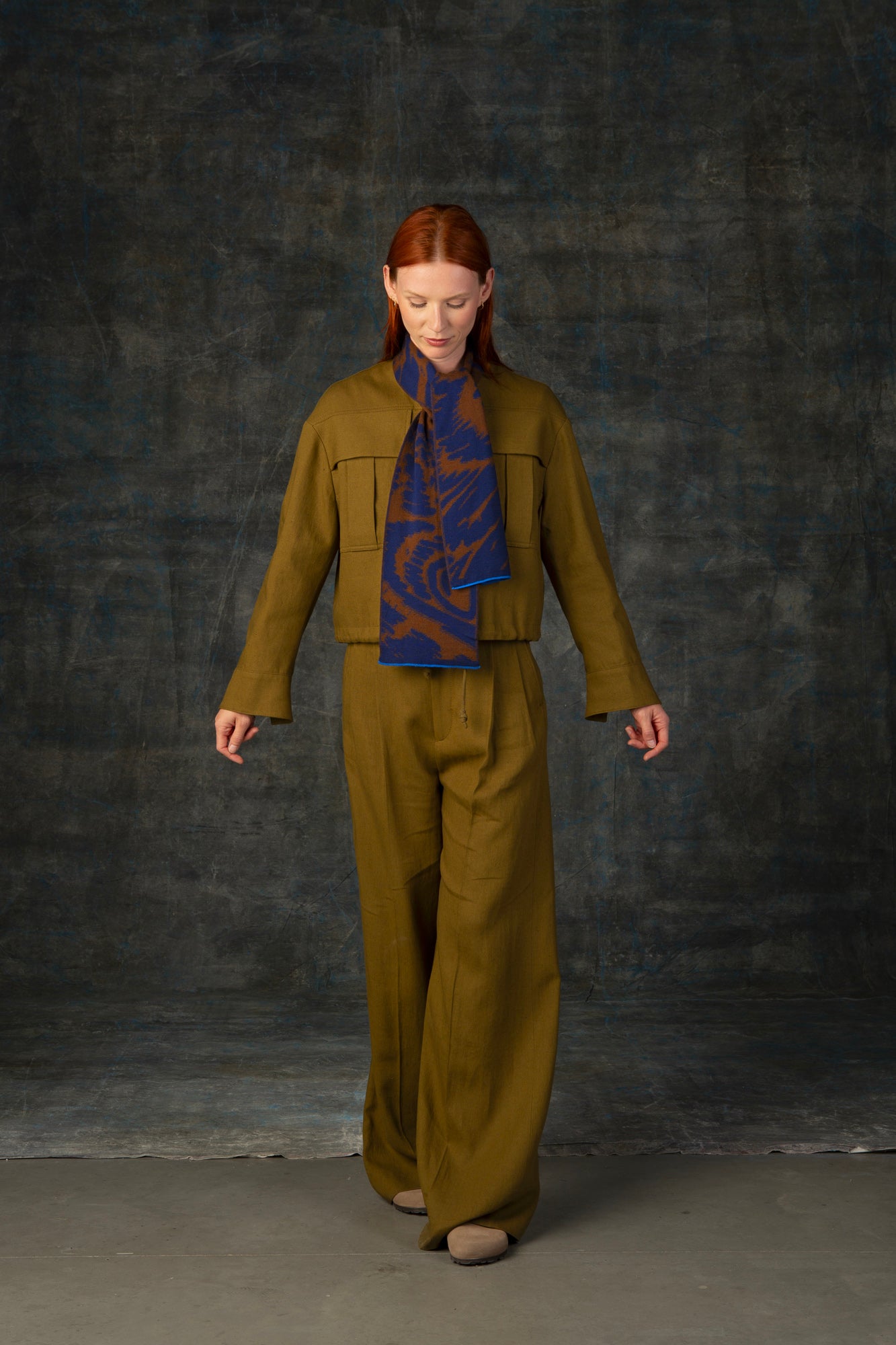 Female model wearing an olive green outfit with a brown and blue mini scarf, with an organic pattern, loosely tied around her neck.