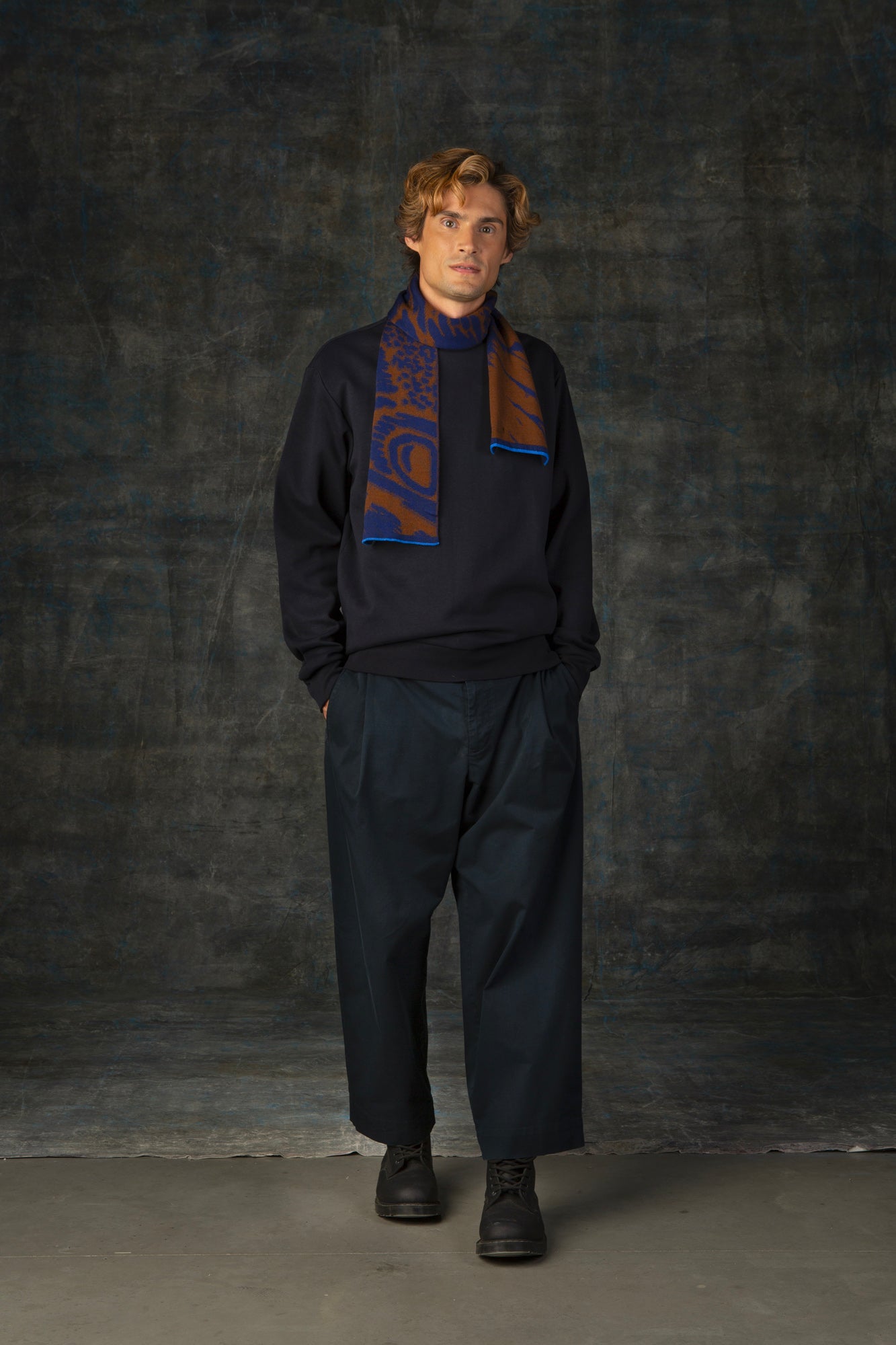 Male model wearing a dark blue outfit with a brown and blue mini scarf, with an organic pattern, draped around his neck.