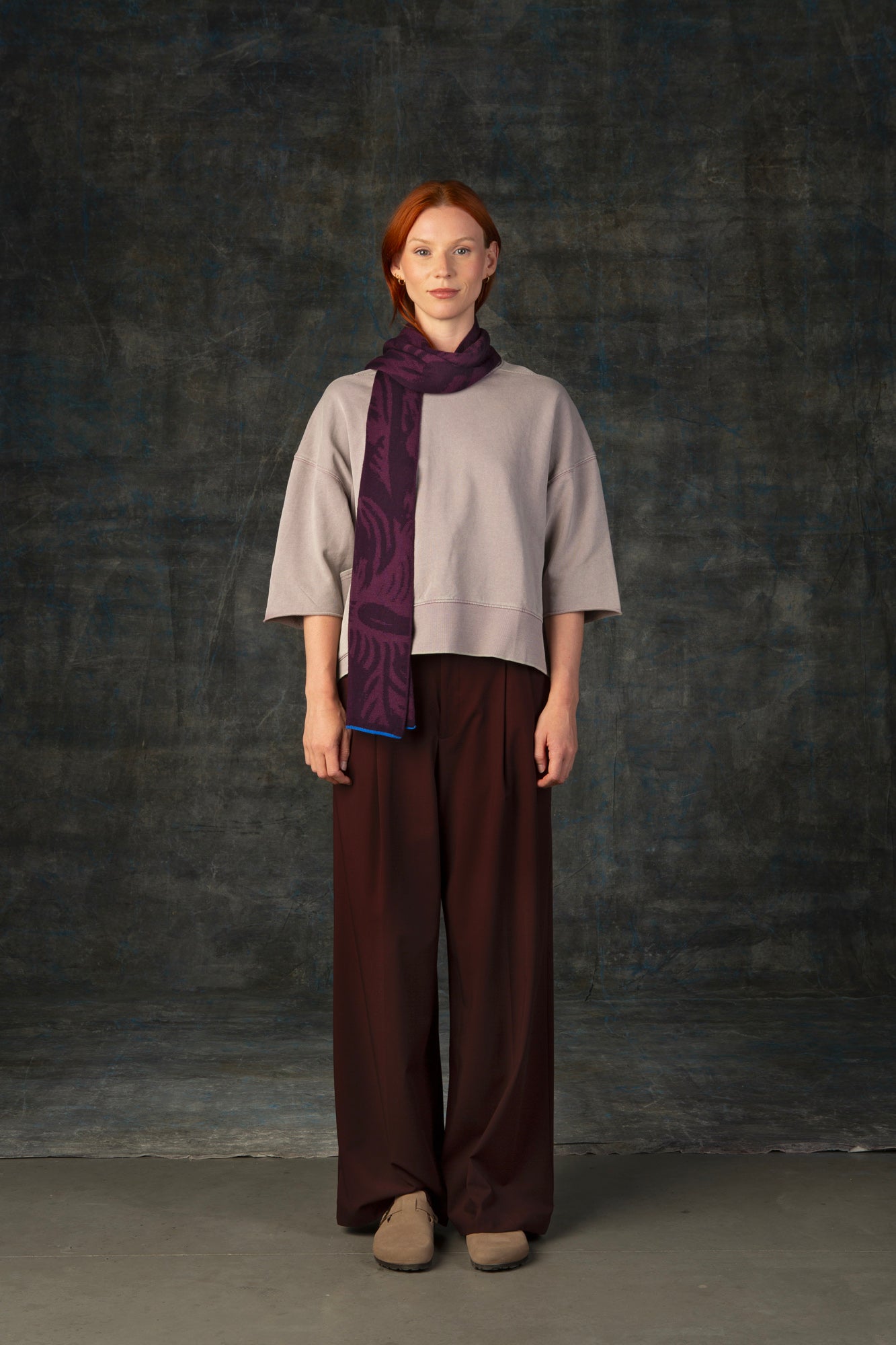 Female model wearing a neutral outfit with a dark purple midi scarf, with nature-inspired pattern draped around her neck, against a dark background.