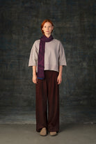 Female model wearing a neutral outfit with a dark purple midi scarf, with nature-inspired pattern draped around her neck, against a dark background.