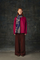 Female model wearing a fuchsia outfit with a dark blue and beige midi scarf draped around her neck, against a dark background.