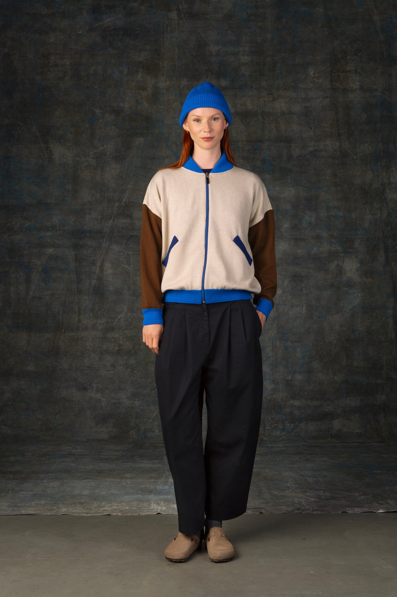 Female model  wearing an ecru and brown knit bomber jacket with cobalt blue details and matching cobalt blue beanie. 