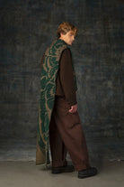 Male model wearing a brown outfit with a green and olive maxi scarf, with an organic pattern, draped over his shoulder.