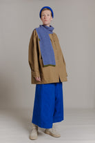 Full length shot of a woman wearing a small knit scarf and matching beanie. She's standing in front of a neutral grey background and is wearing a brown coat  and wide blue trousers. The knit scarf has a checkered pattern in cobalt blue and beige with olive green details.
